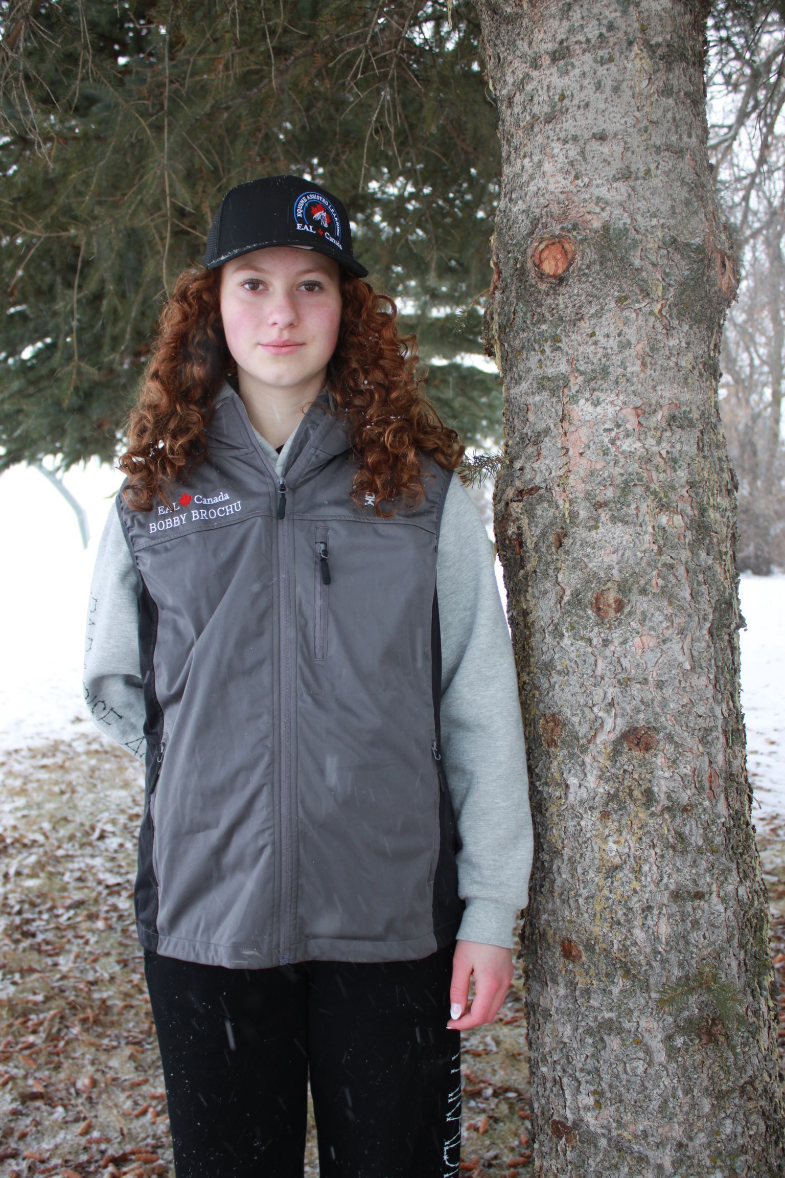 Person with curly hair wearing a vest and hat, standing near a tree in a snowy environment.