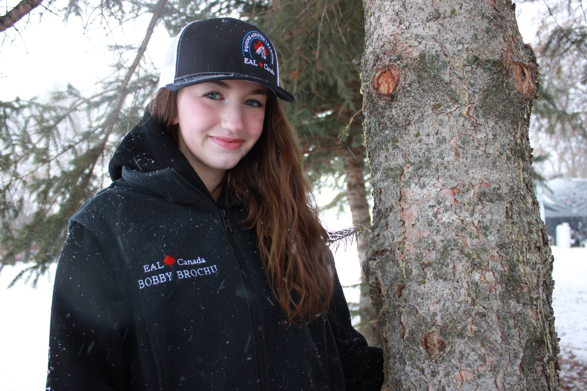 Woman in black hat and hoodie smiles next to a tree in a snowy outdoor setting.