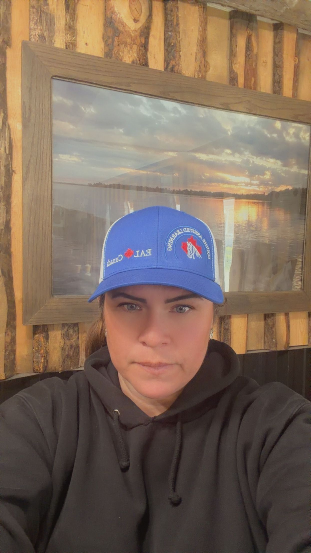 Person in a blue cap and black hoodie taking a selfie in front of a framed scenic wall photo.