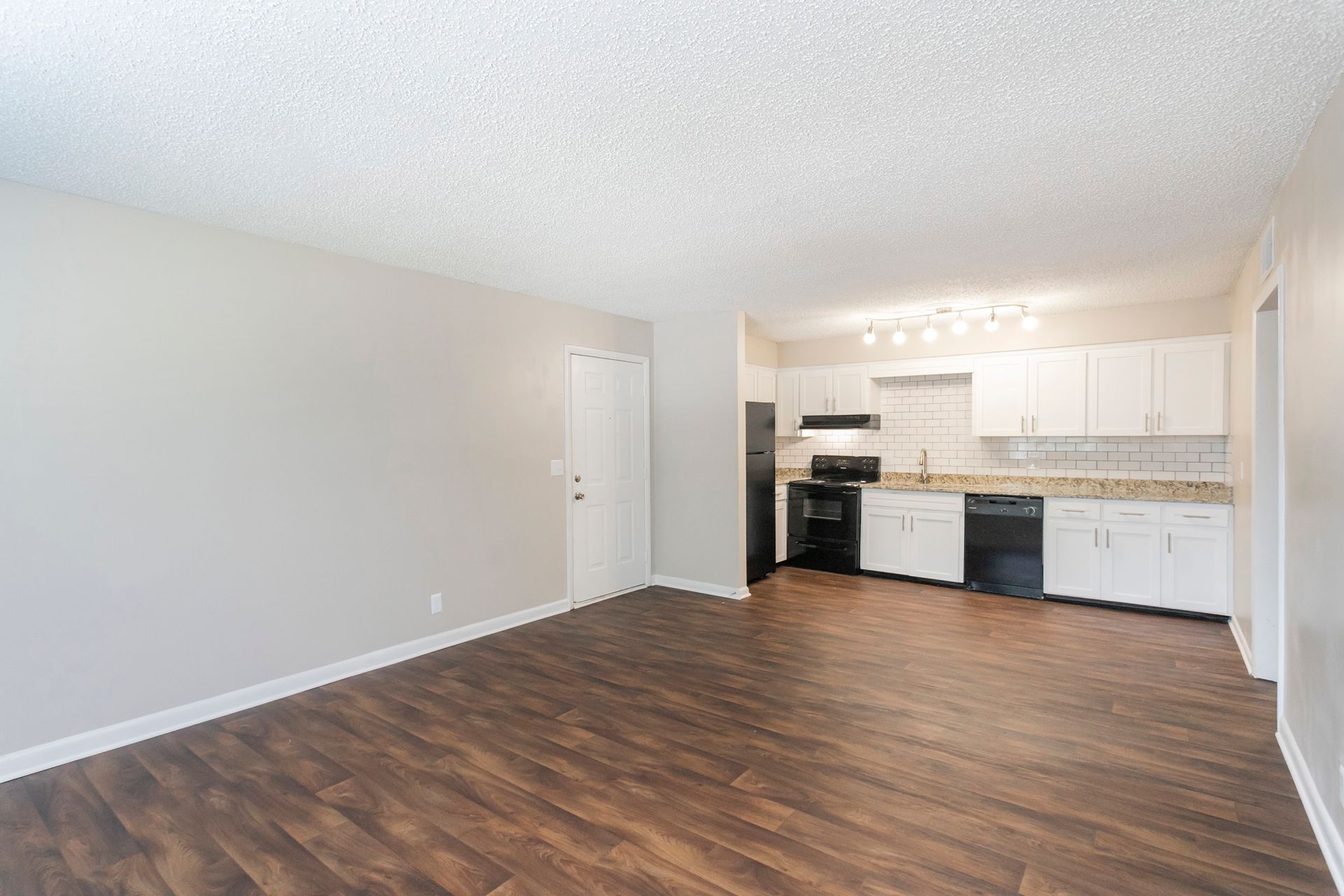 A spacious, open-concept room with wood-look flooring, light walls, a white entry door, and a modern kitchen area.