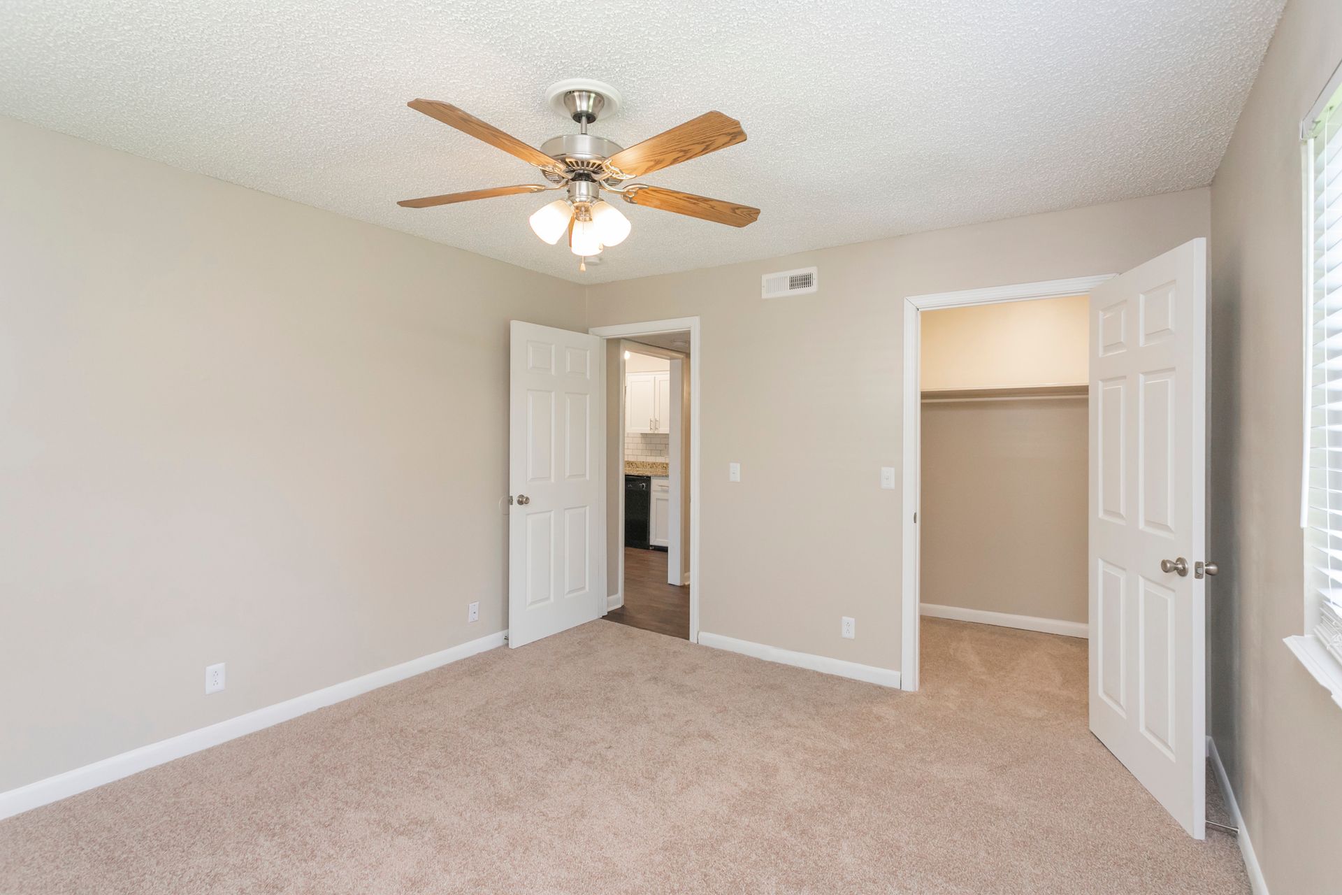 A spacious bedroom with beige walls, tan carpeting, a ceiling fan, a doorway to another room, and an open closet.
