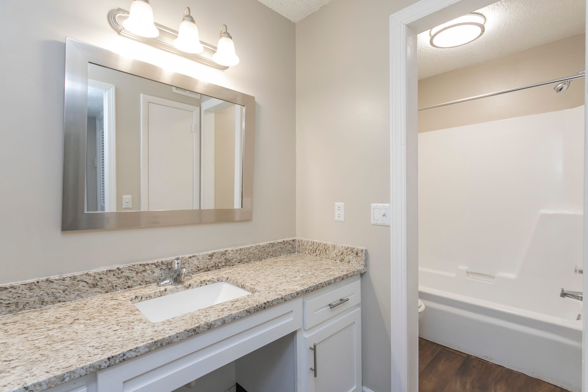 A bathroom with a white vanity, granite countertop, rectangular sink, mirror, vanity lights, and a shower-tub combo.