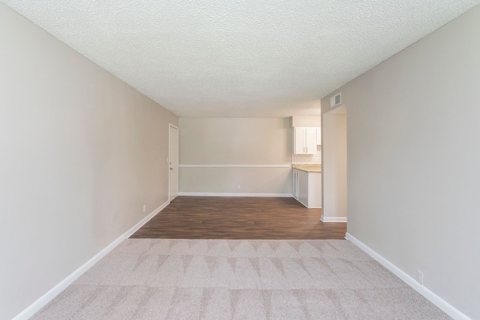 An empty apartment living area with beige walls, a gray carpeted foreground, and a wood-floored dining space in the back.