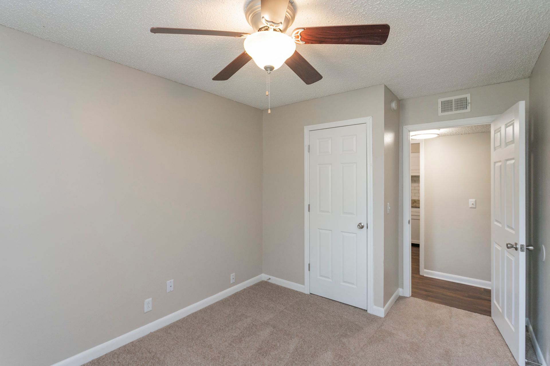 A small, empty bedroom with beige walls, tan carpeting, a ceiling fan, and two white doors.