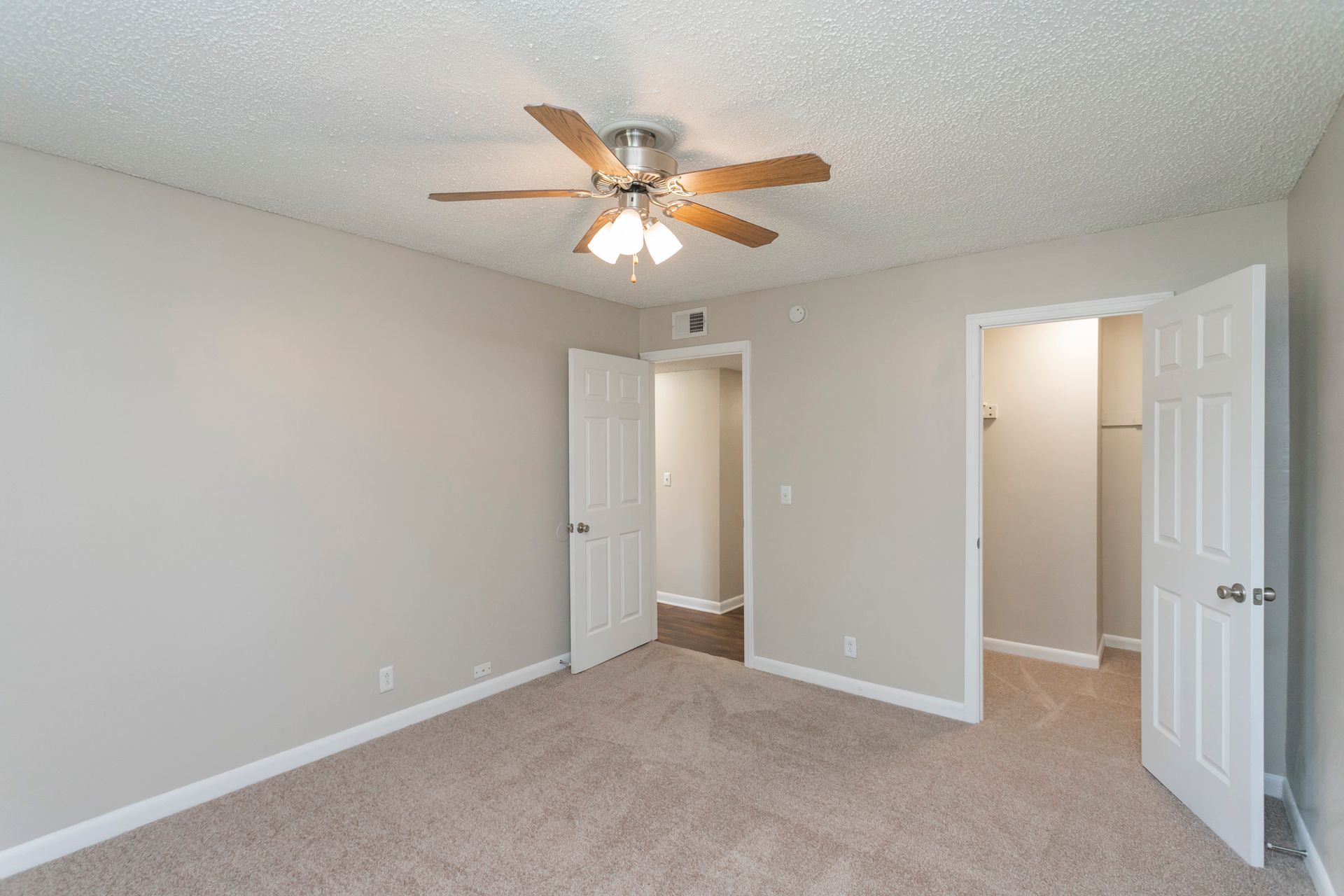 Empty bedroom with light grey walls, carpeted floor, ceiling fan, and open doorways to a hallway and a closet.