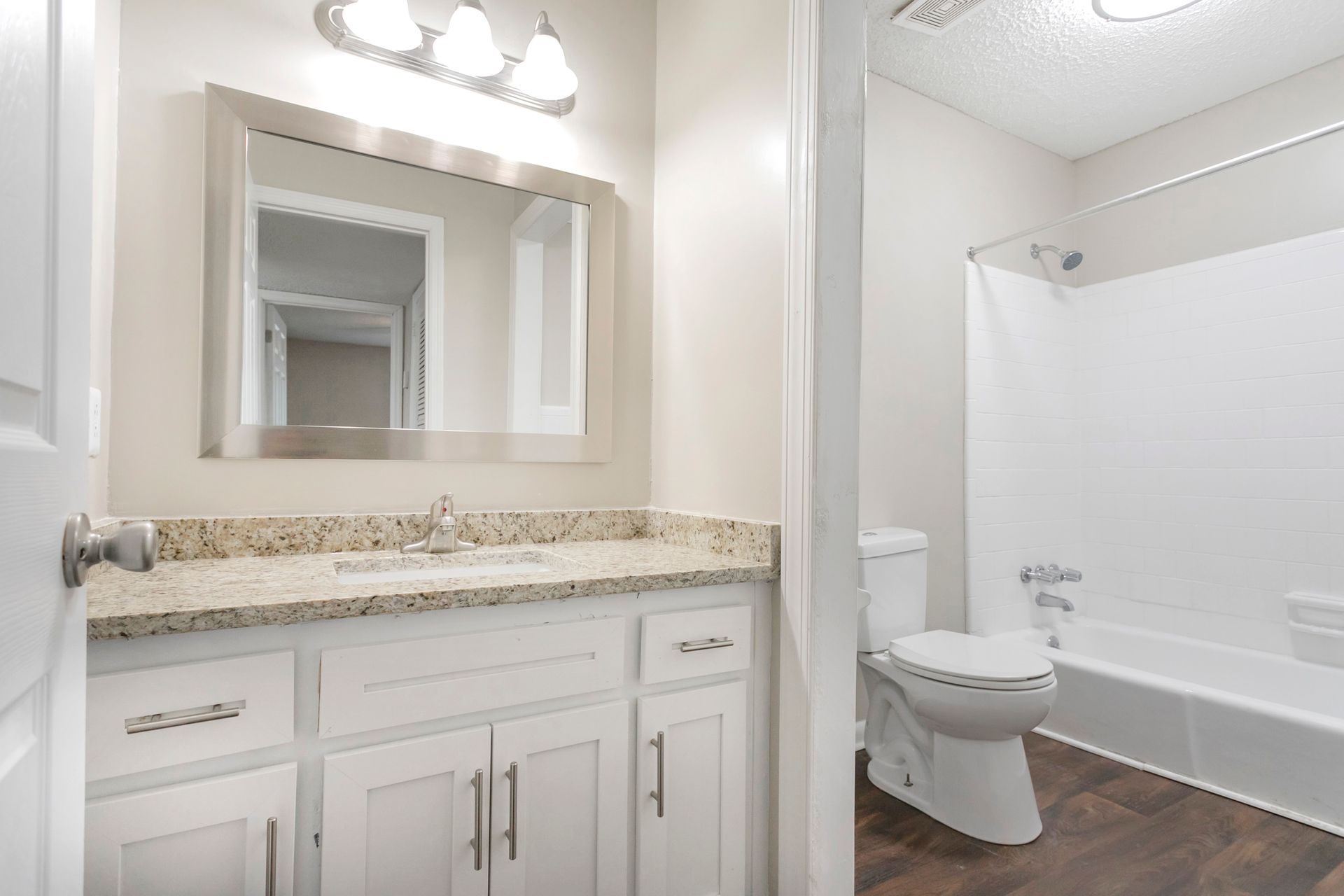 A bathroom featuring a white vanity with a granite countertop, a rectangular mirror, a toilet, and a bathtub with shower.