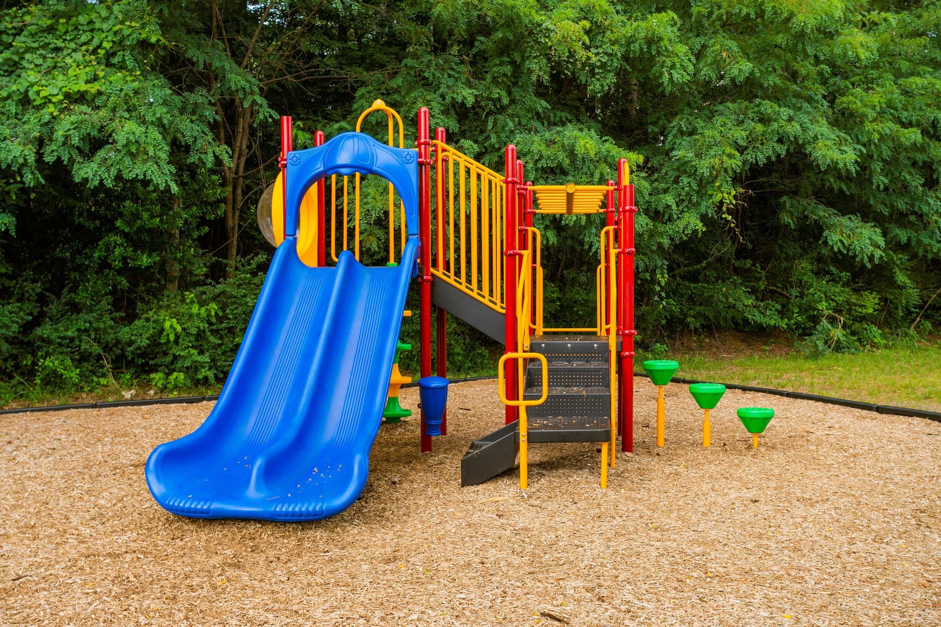 A colorful playground slide set with red posts, yellow railings, and blue slides surrounded by wood chips and trees.