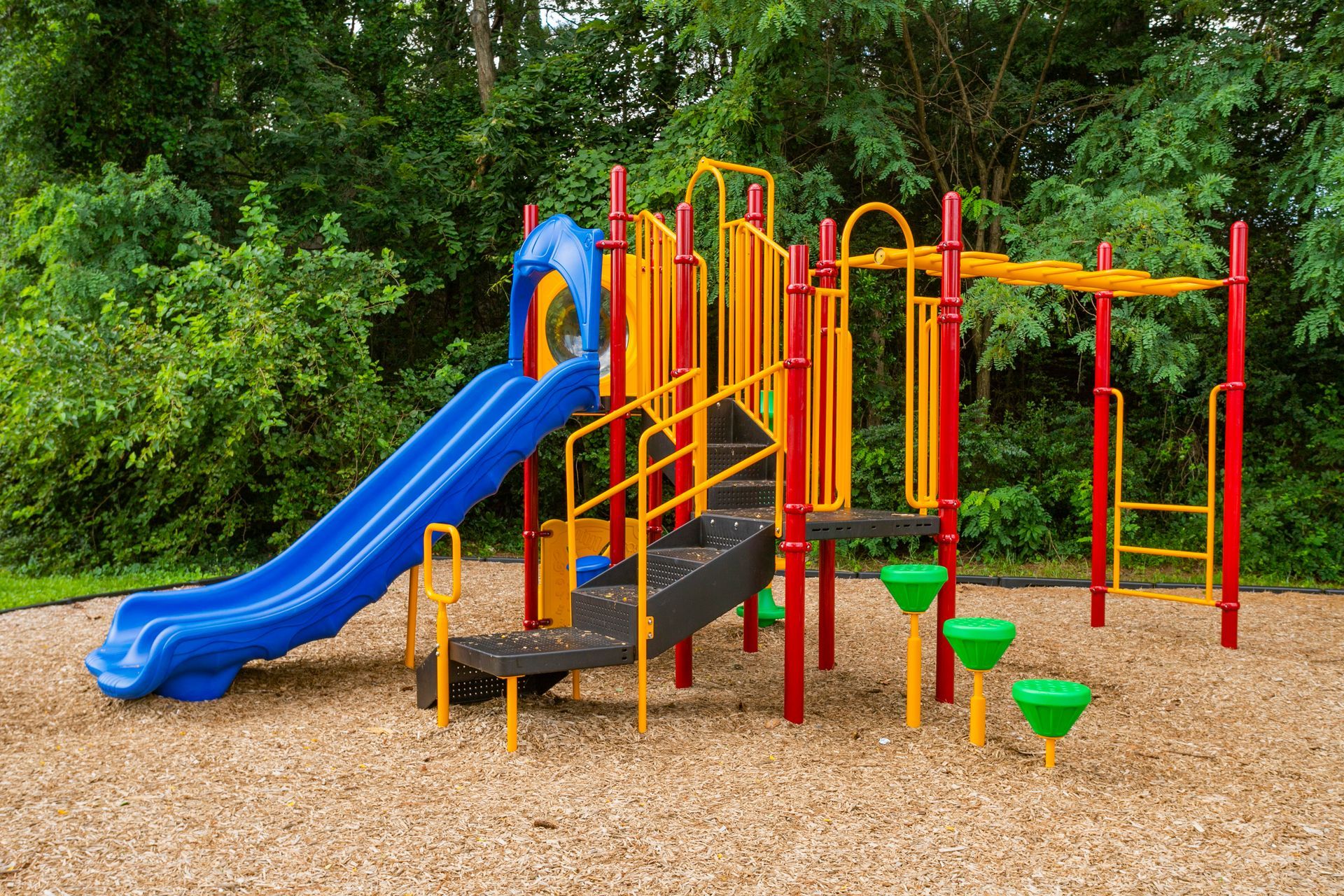 A colorful playground structure with a blue slide, yellow railings, and green stepping pads set on wood chips.