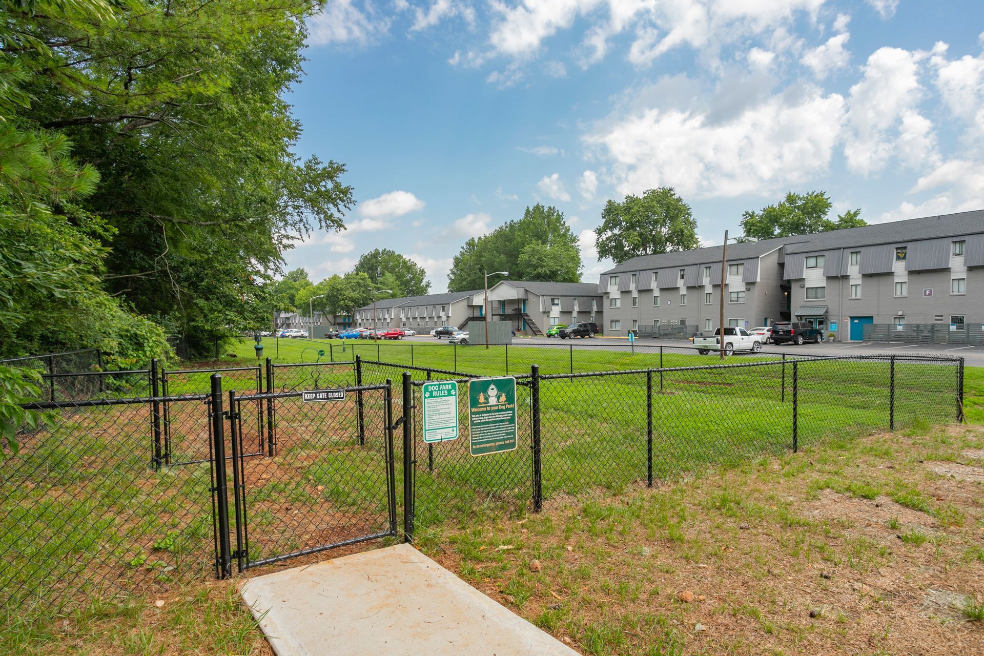 A fenced-in dog park with a gate and a concrete entrance path, situated in front of a multi-story gray apartment complex.