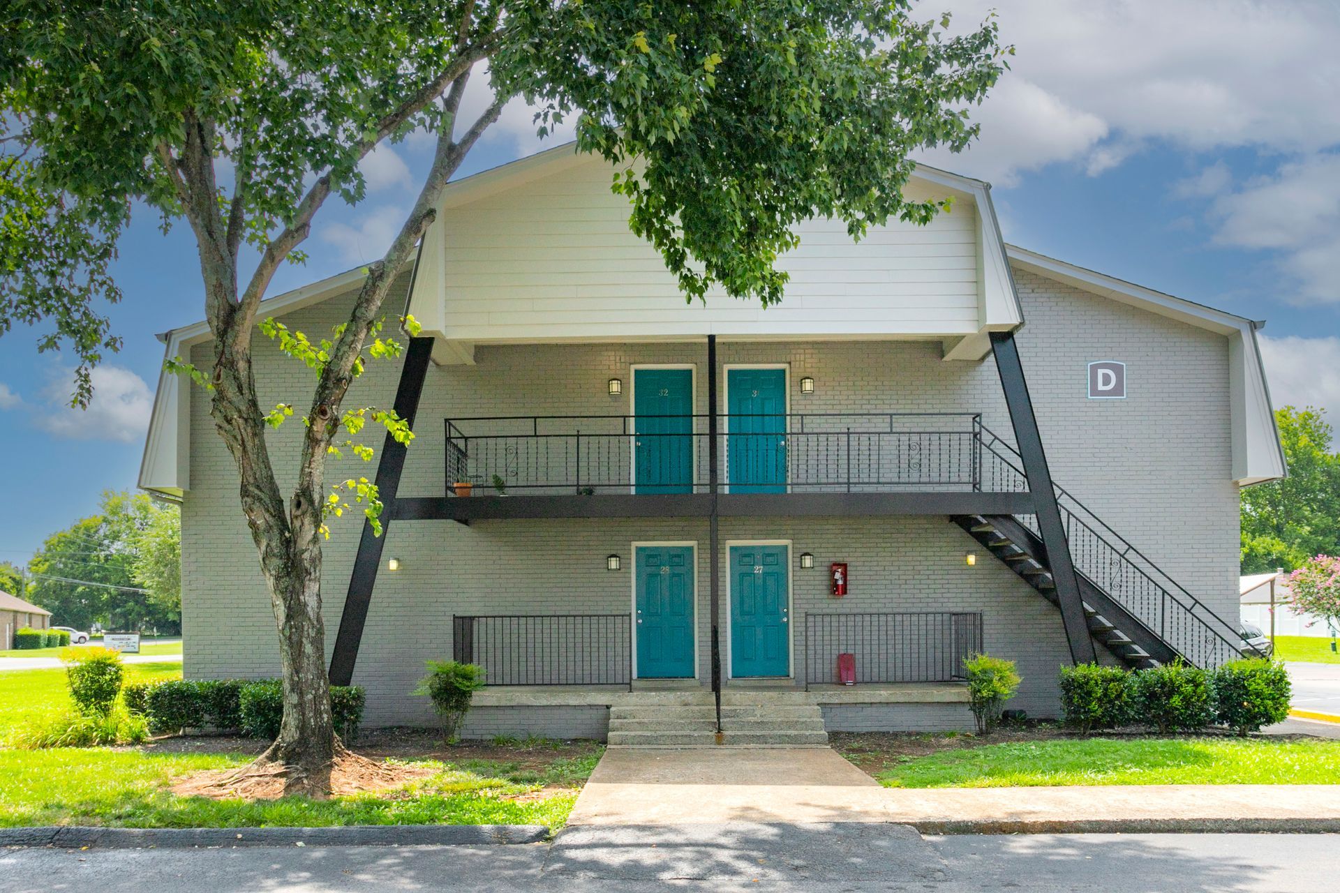 A two-story apartment building with light gray siding, teal doors, and a black metal staircase, framed by a large tree.