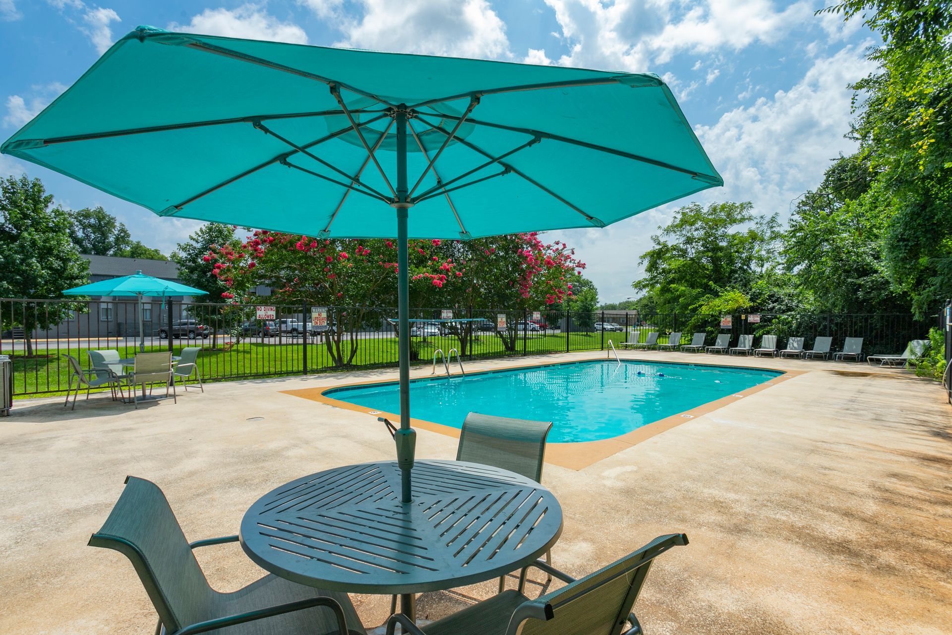 A round patio table with chairs under a teal umbrella beside an outdoor swimming pool on a sunny day.