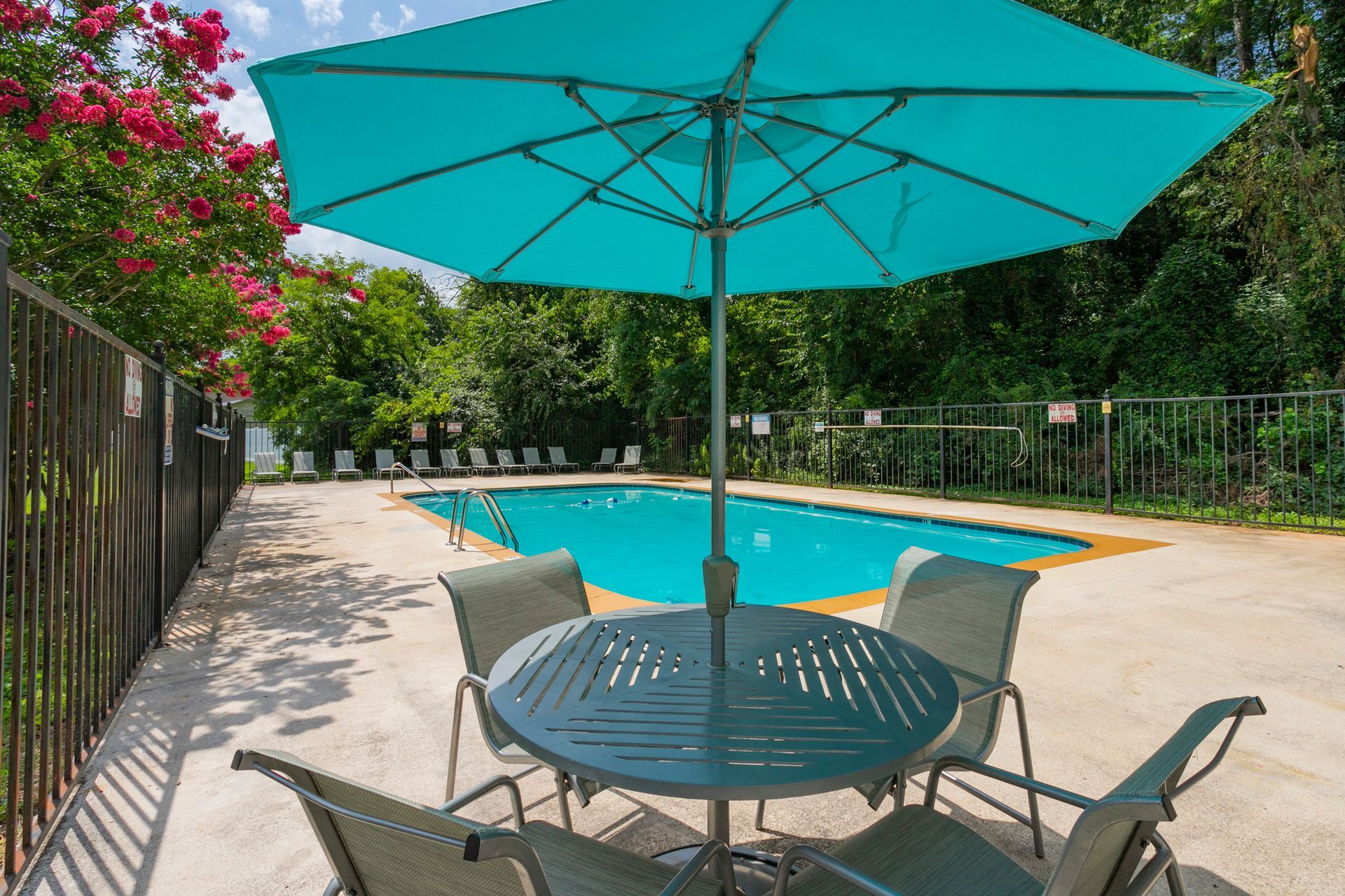 A turquoise umbrella shades a round table with four chairs on a pool deck next to a bright blue swimming pool.