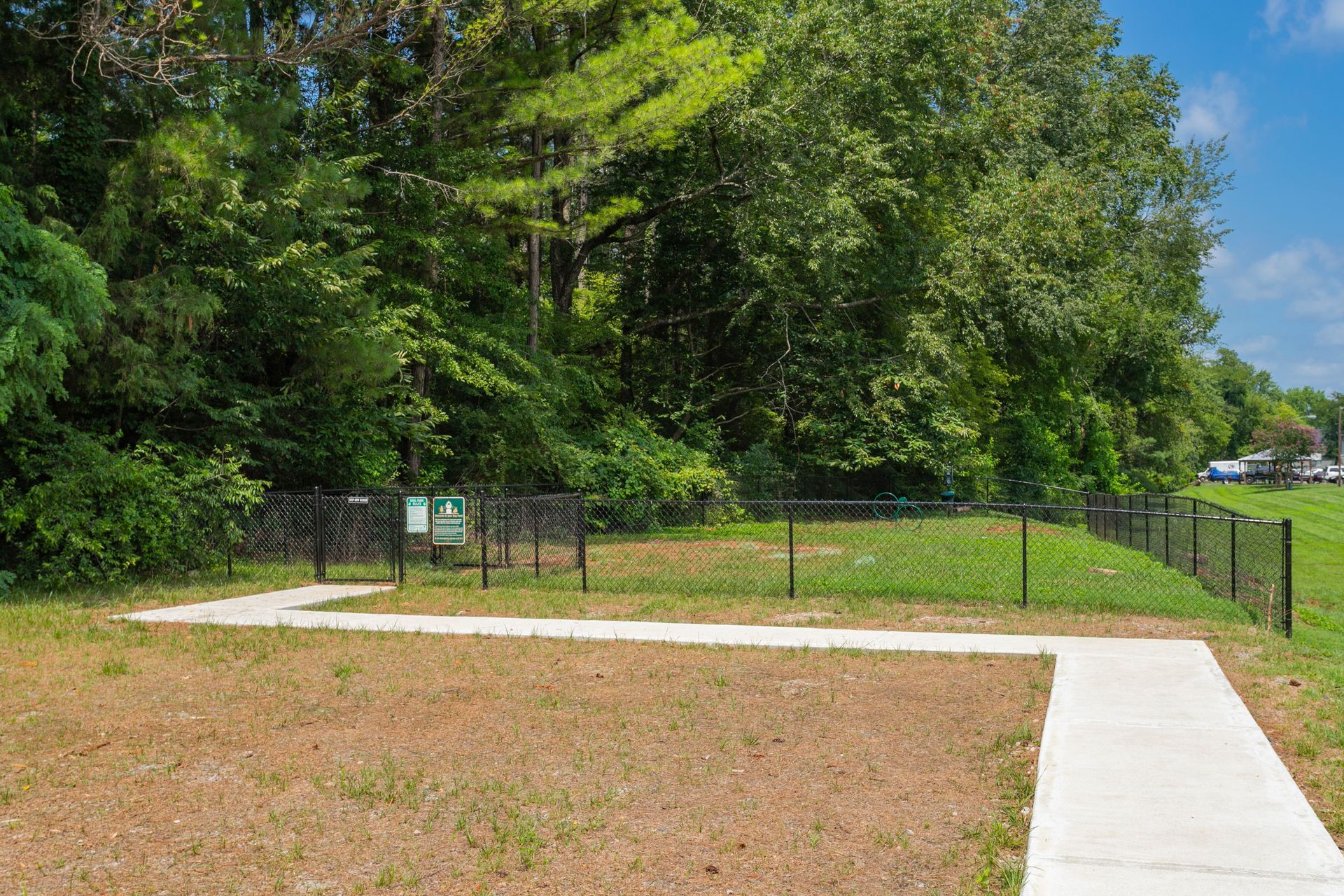 A concrete sidewalk leads to a fenced-in dog park area bordered by a dense line of trees on a sunny day.