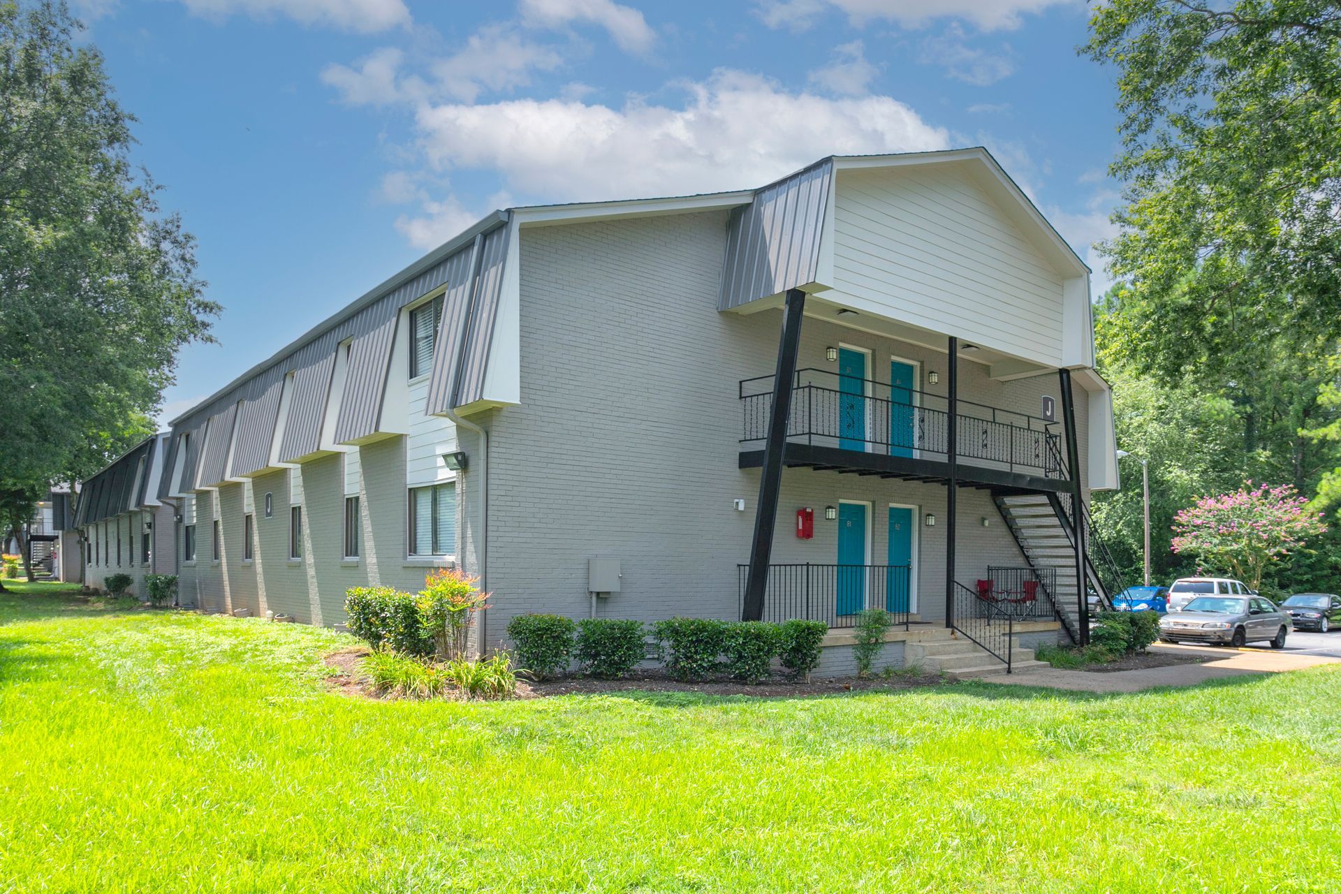 A multi-story gray apartment building with a slanted roof, external stairs, and bright teal doors, set in a grassy lawn.