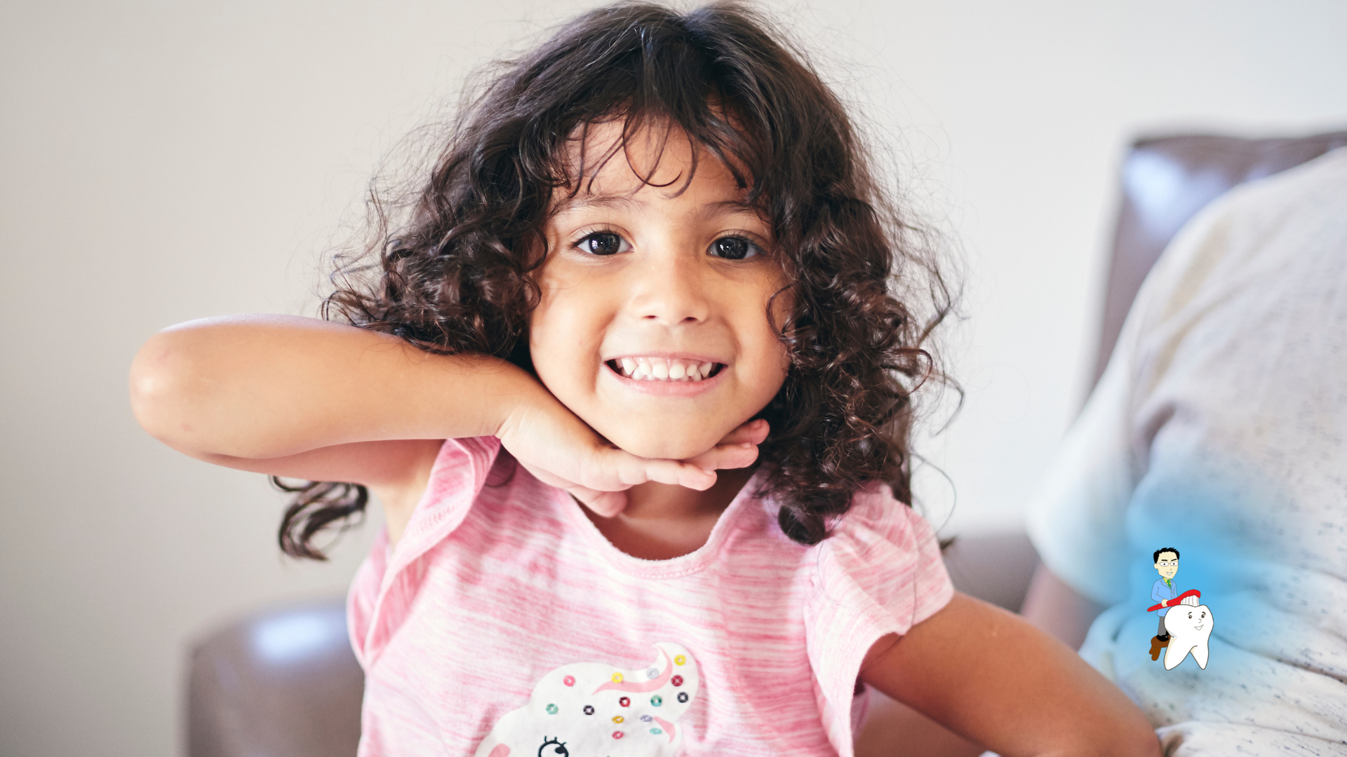 A little girl with curly hair is sitting on a couch and smiling.