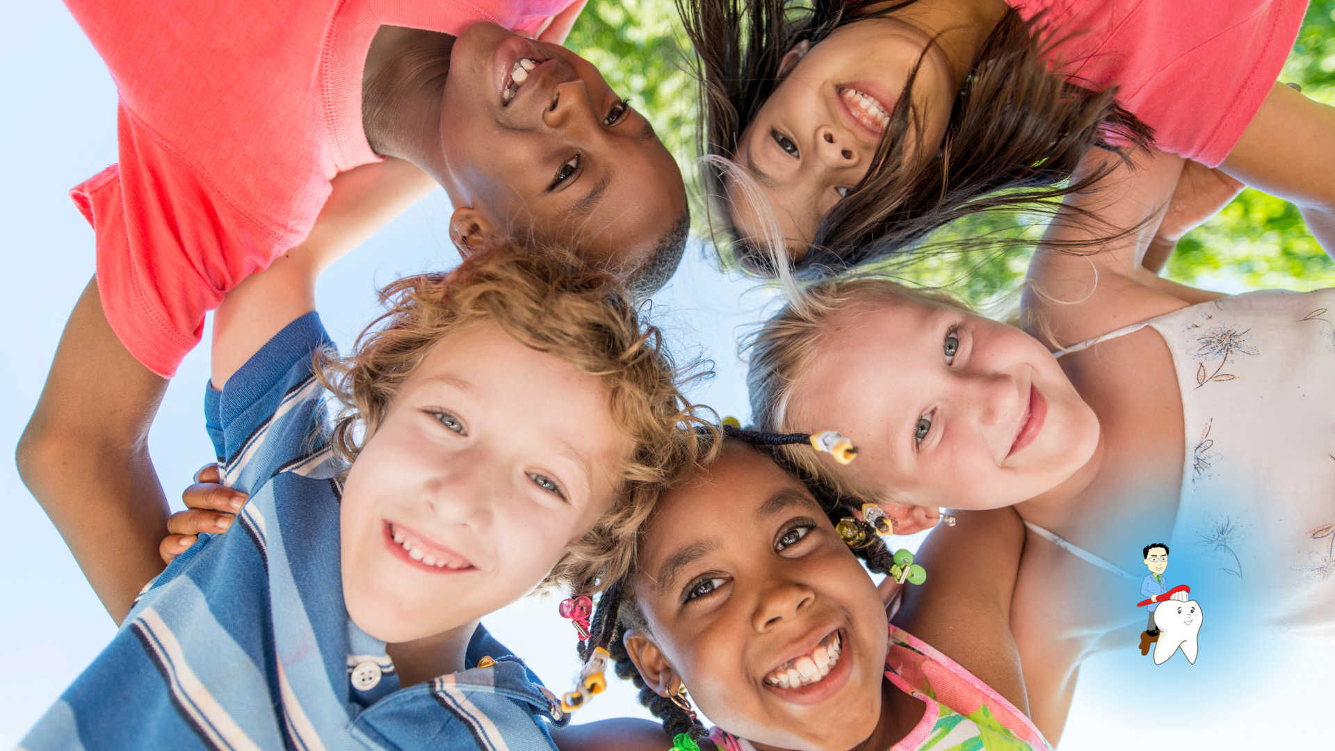 Five smiling children leaning in a circle, looking down at the camera outdoors.