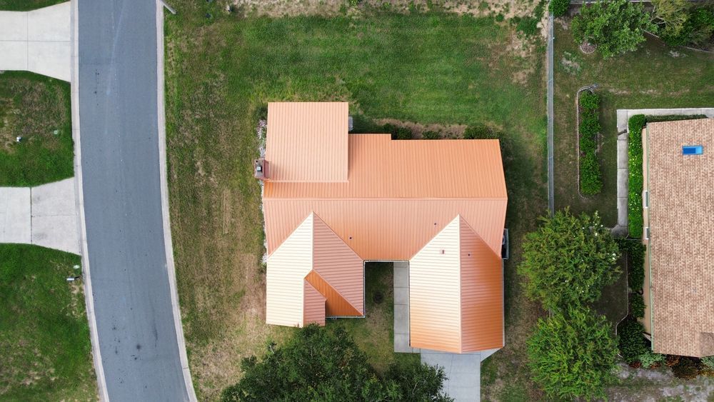 Aerial view of a tan-roofed house beside a curved road, with grass and neighboring homes nearby
