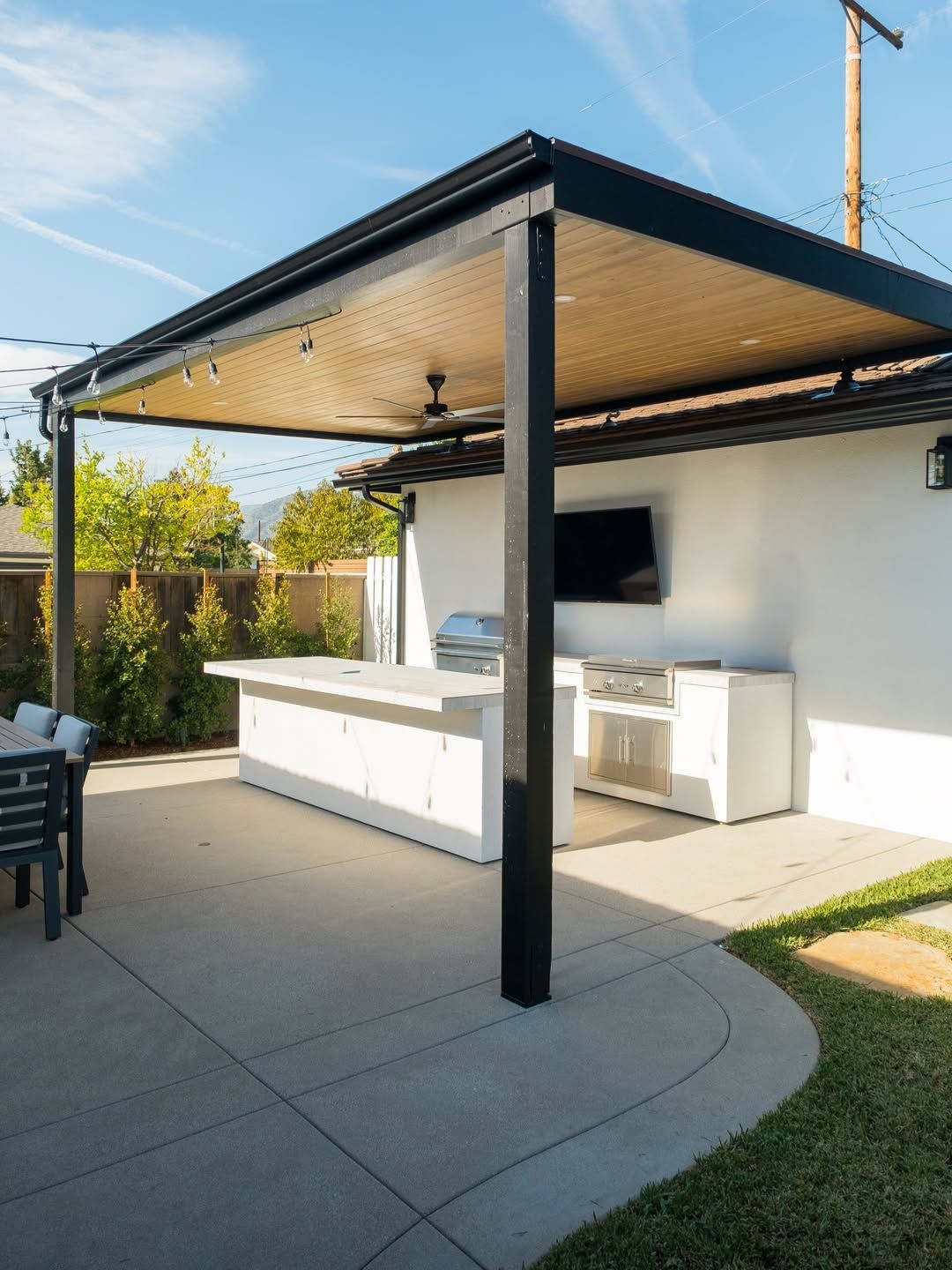 Outdoor kitchen under a black-framed pergola with a concrete counter, grill, and mounted TV.
