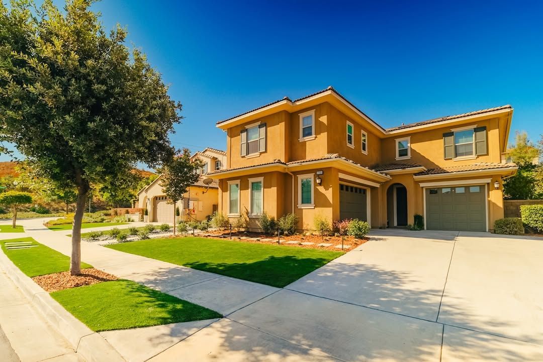 Two-story tan house with a green lawn, sidewalk, and driveway under a blue sky.