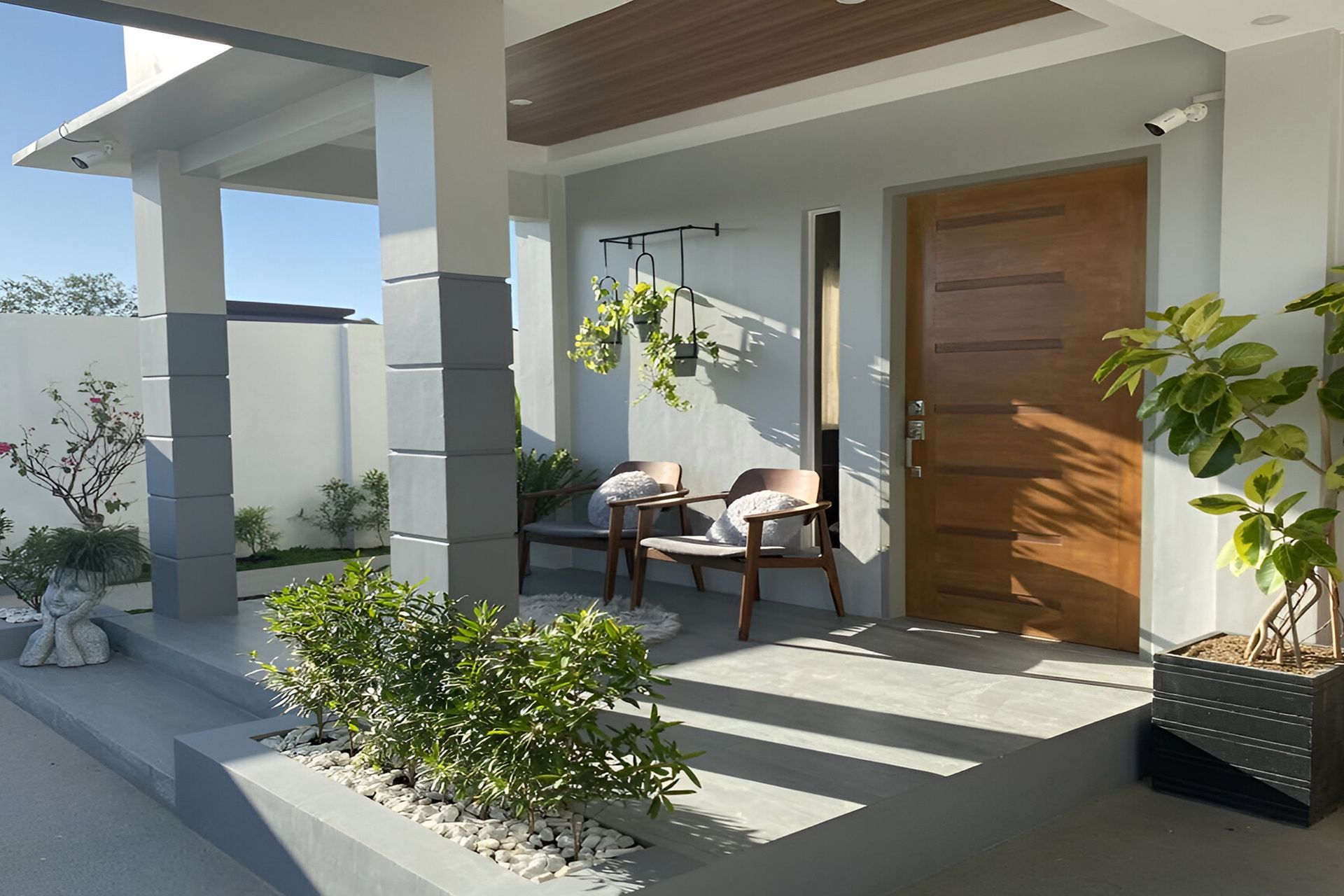 Modern front porch with wood door, chairs, hanging plants, and potted greenery in sunlight