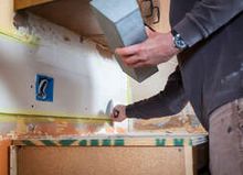 Handy worker drywall installer apply drywall mud with a putty knife above a kitchen counter to a damaged drywall panel. The man is wearing a blue sweater and black watch holding a mud tray in his left hand.