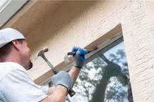 Maple Ridge Window Installer holding a crow bar to a exterior window. The worker is preparing to hit the crow bar with a hammer to remove the window for a retrofit window replacement and installation. The worker is wearing a white backwards hat and white T-shirt with blue and grey gloves.