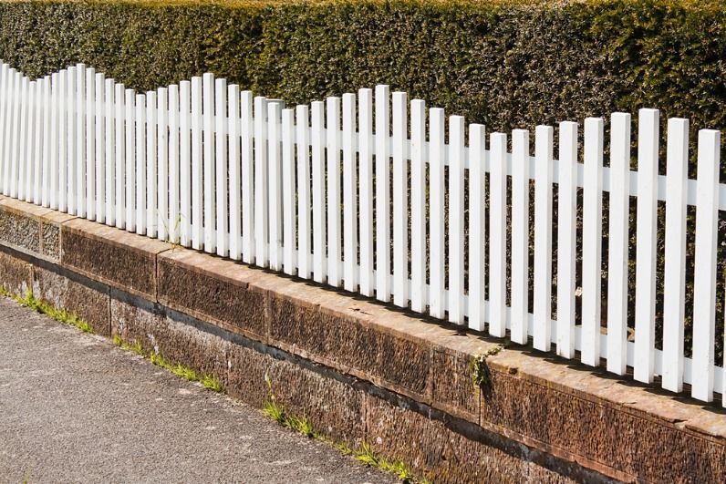 A White Picket Fence is Sitting Next to a Stone Wall — Brendan's Landscaping and Fencing In Bundaberg, QLD