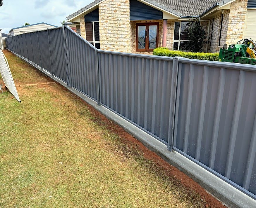A Green Fence With Brick Pillars is Surrounded by Trees and Bushes — Brendan's Landscaping and Fencing In Bundaberg, QLD