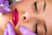 Gloved hands applying bright pink lip color to a closed-eyed face with a needle-like tool