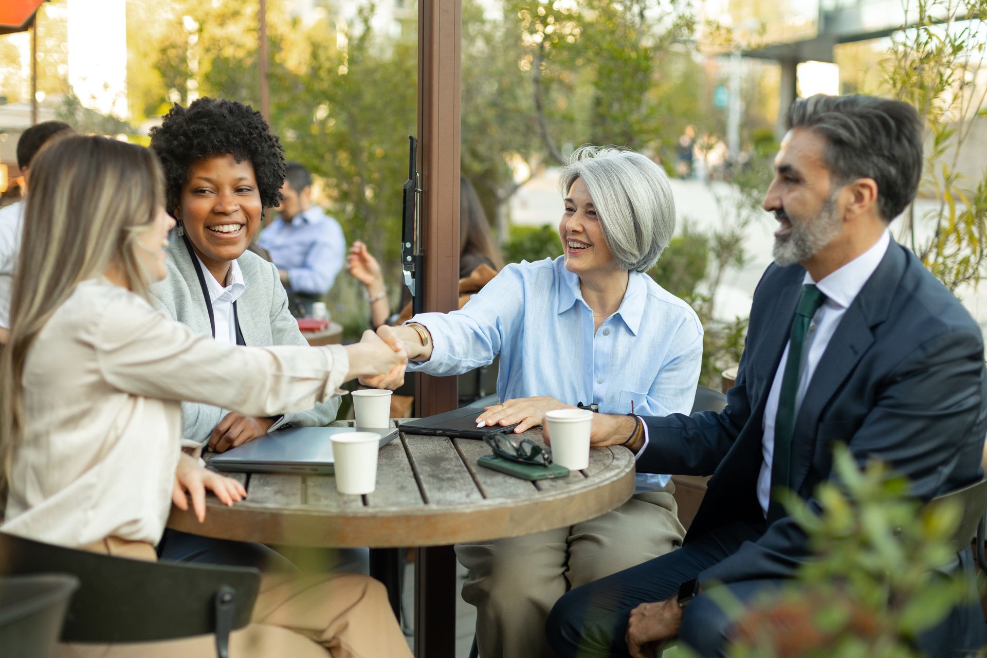 Four people chatting and shaking hands at an outdoor café table with coffee cups