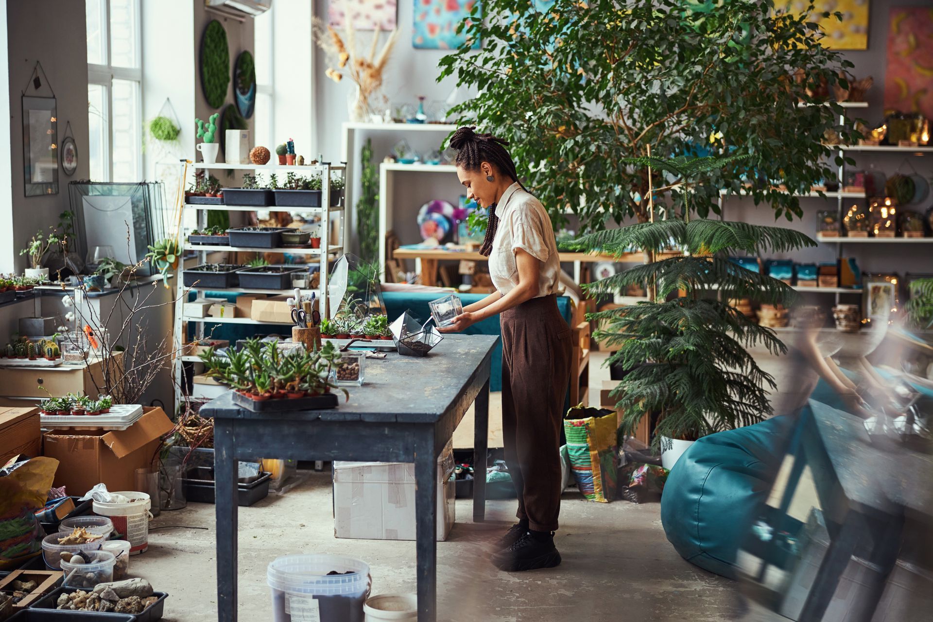Person arranging plants at a workbench in a bright, cluttered plant studio with shelves and greenery