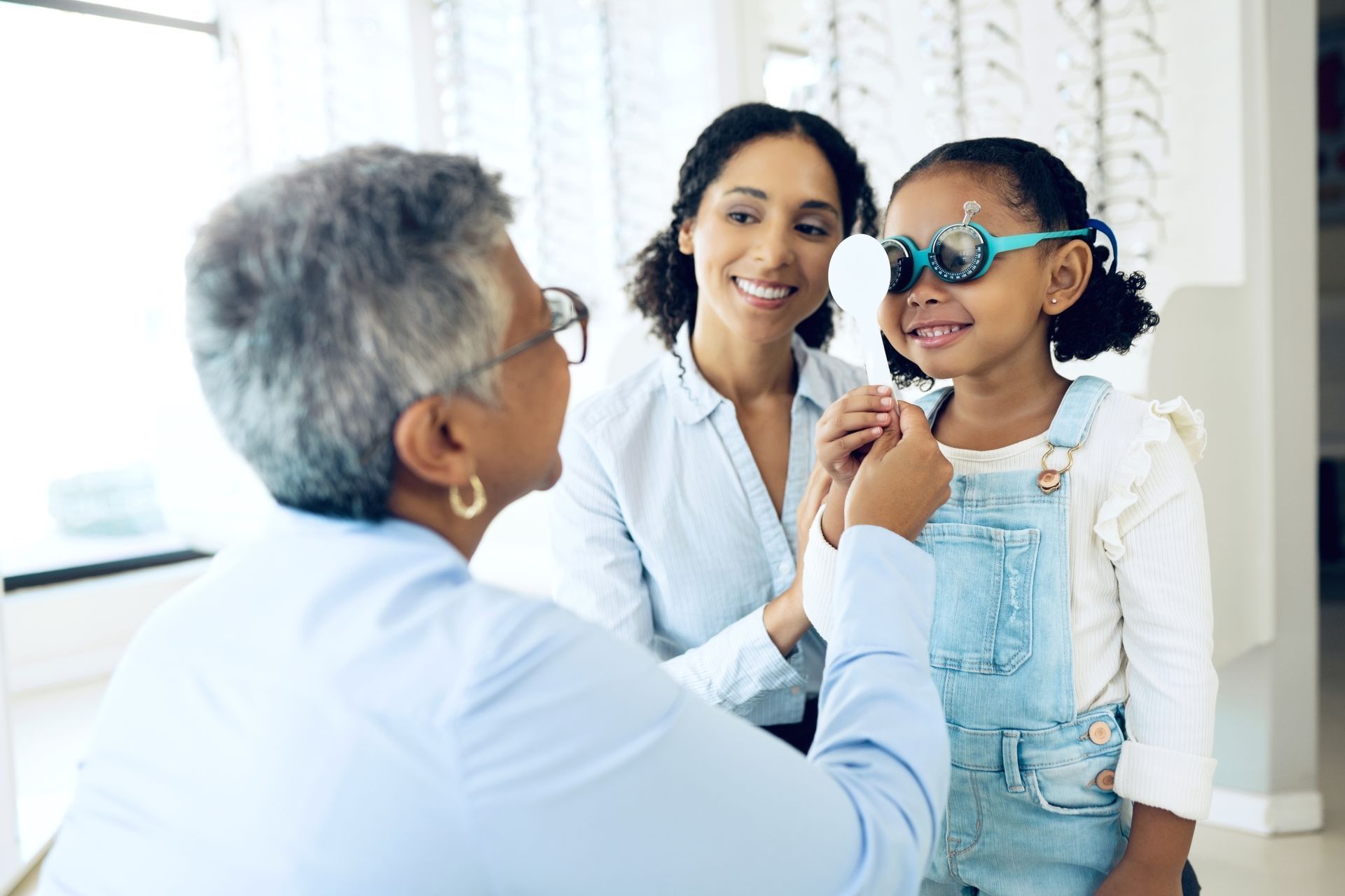 Doctor examining a smiling child wearing glasses while a caregiver watches in a bright clinic.