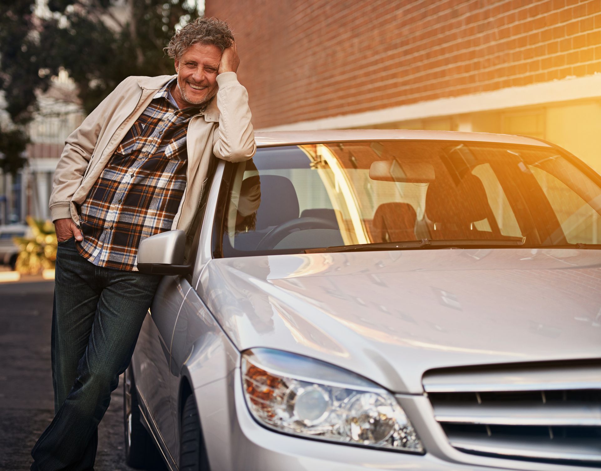 Smiling man leans on silver car beside brick wall at sunset