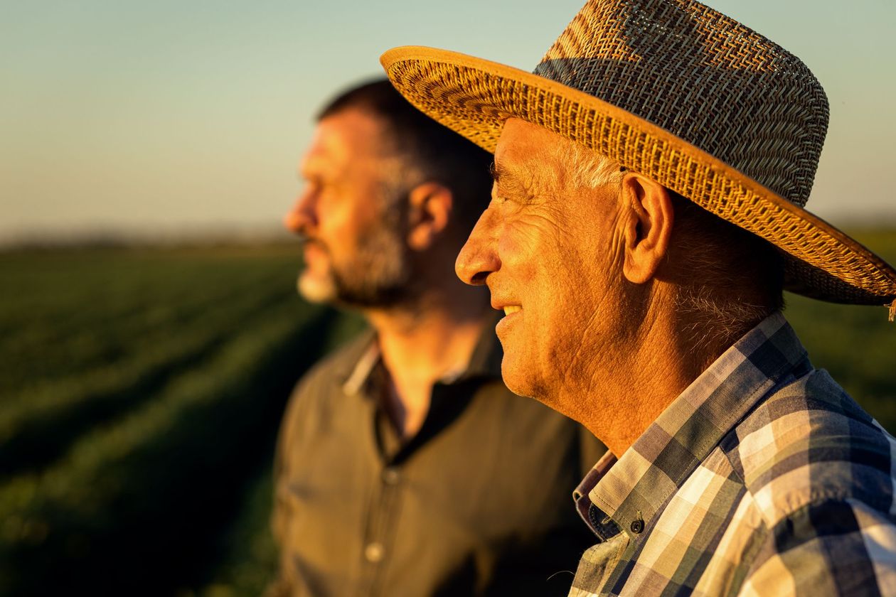Two men in hats stand in a field at sunset, one in sharp focus and smiling in the foreground.
