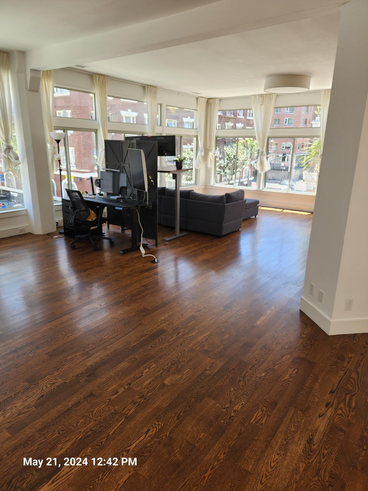 A bright, open-plan living space with polished dark wood floors, a desk setup, and a gray sofa by large windows.