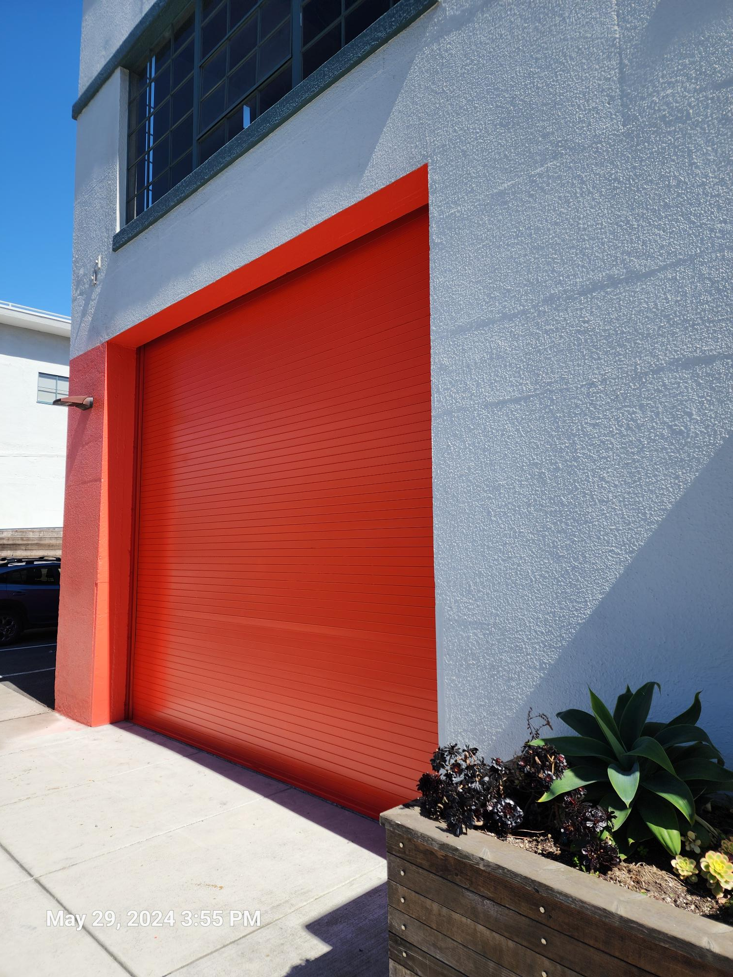 A bright orange, slatted rolling garage door is set into a textured, white-painted exterior wall next to a planter box.