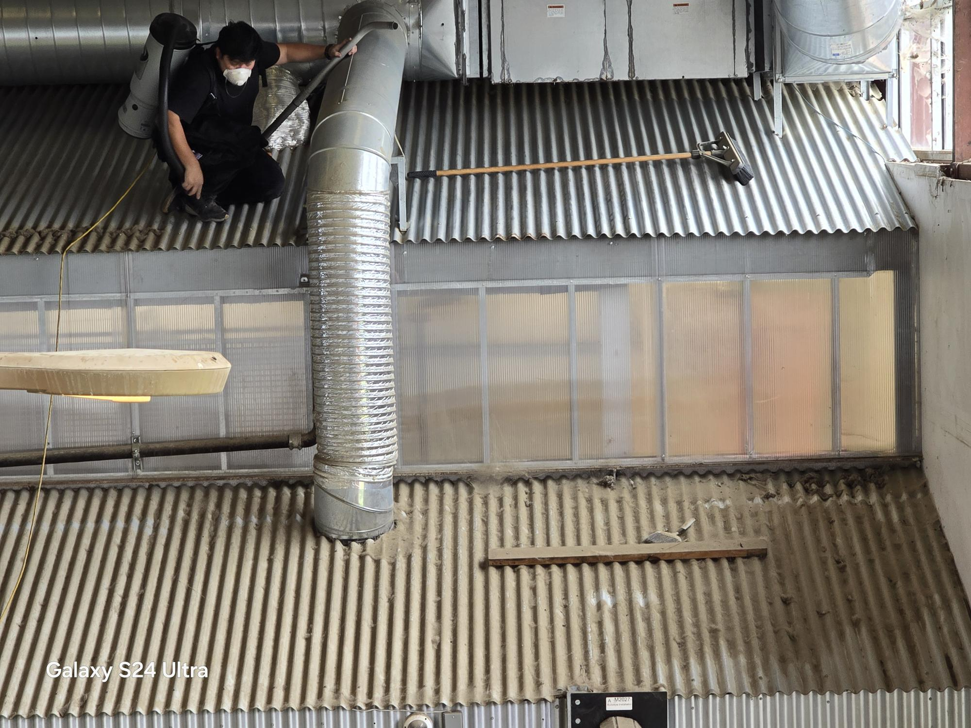 A worker in a mask and black uniform cleans a corrugated metal roof near industrial ventilation ducts.