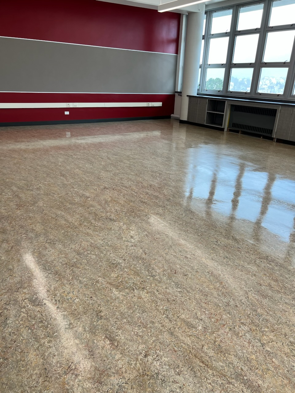 An empty classroom with a polished, speckled floor, a red and grey wall, and a large window with white frames.