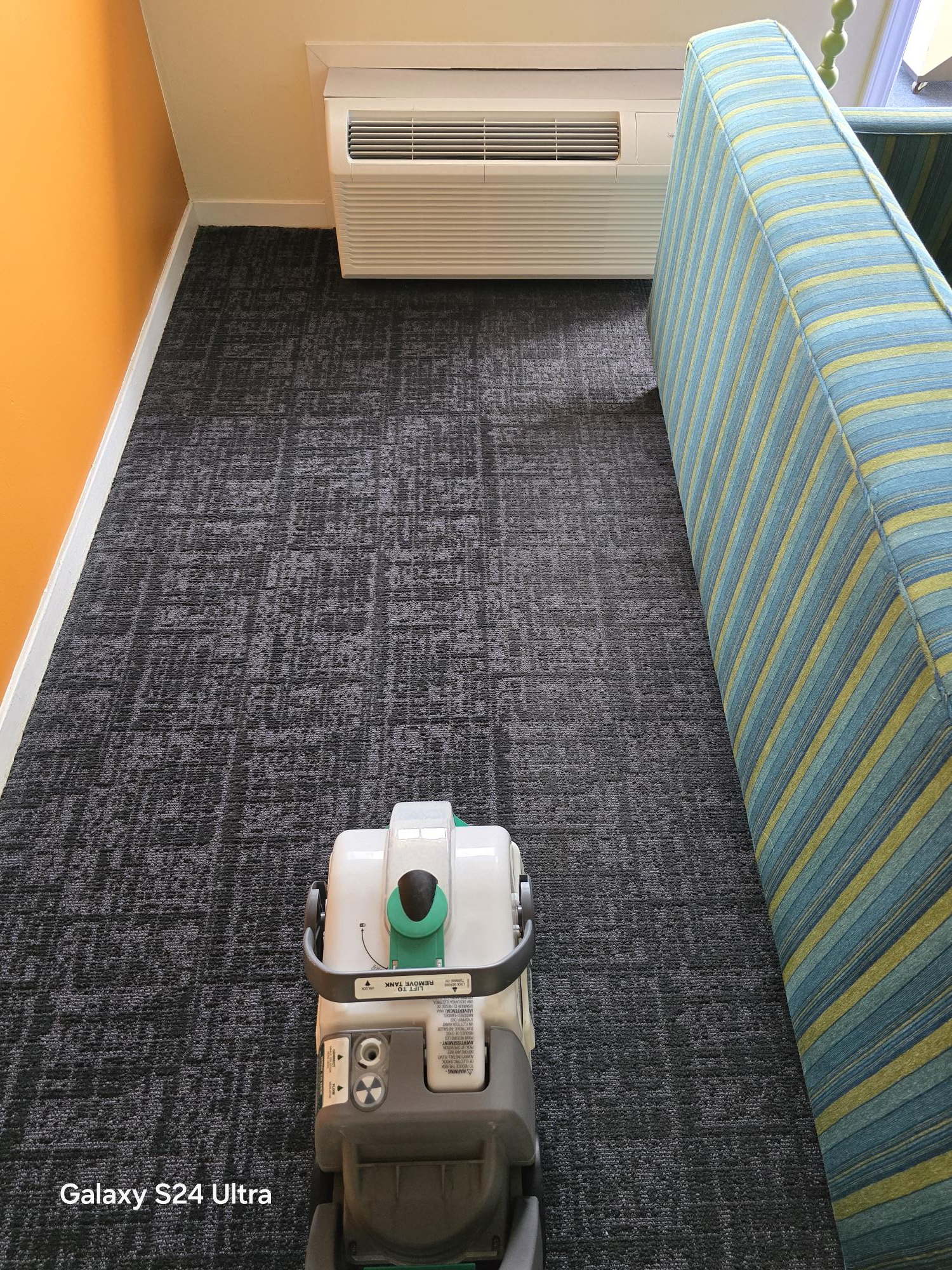 A commercial carpet cleaning machine operates on a dark, patterned carpet next to a blue-striped sofa in a room.