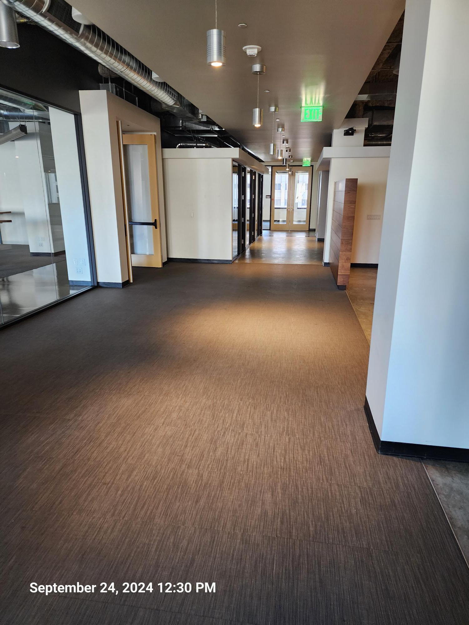 A wide, empty office hallway with grey carpet, white walls, and a row of hanging pendant lights leading to a glass door.