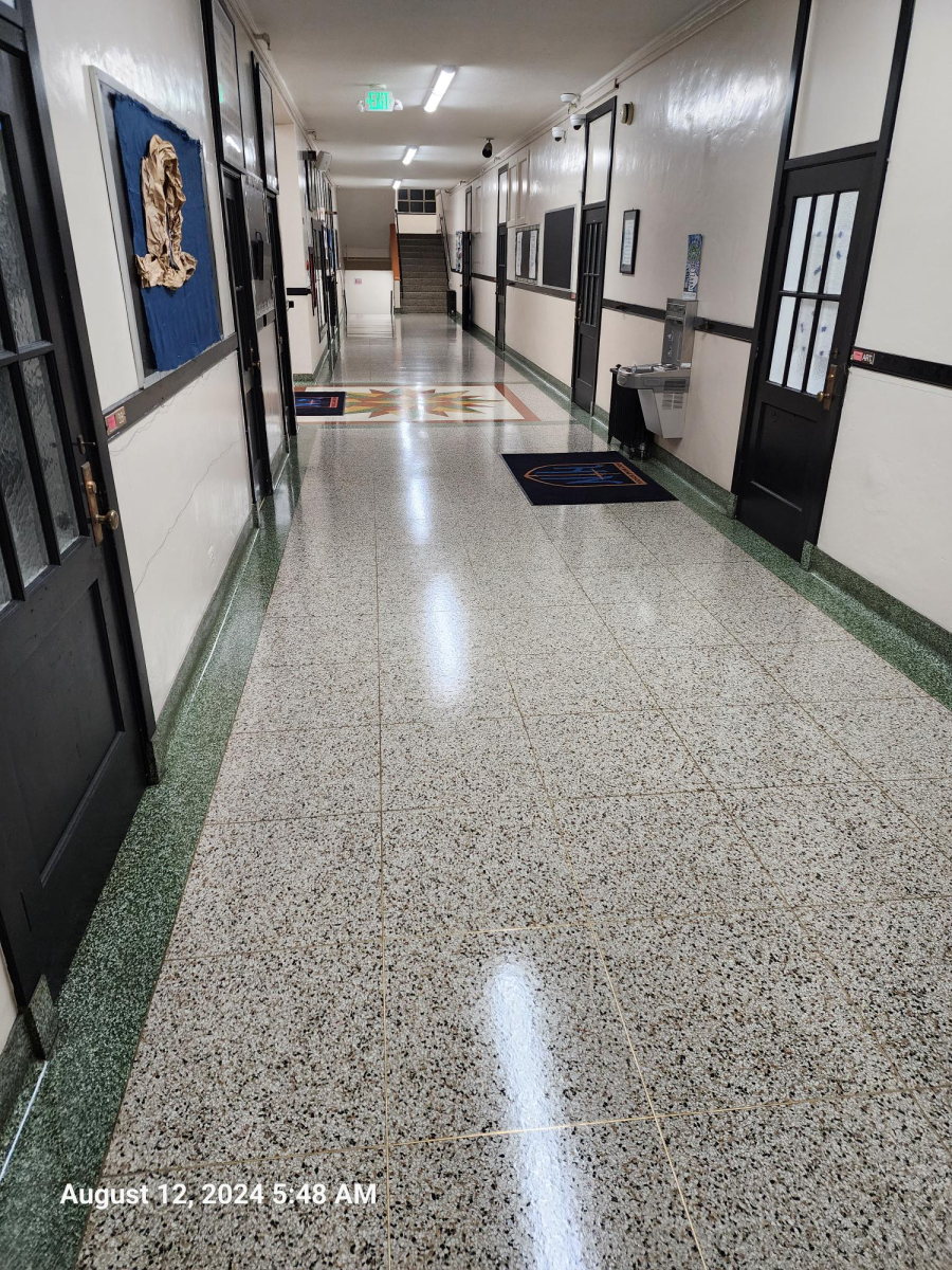 A long, empty school hallway with terrazzo flooring, white walls, dark-framed doors, a blue bulletin board, and exit signs.
