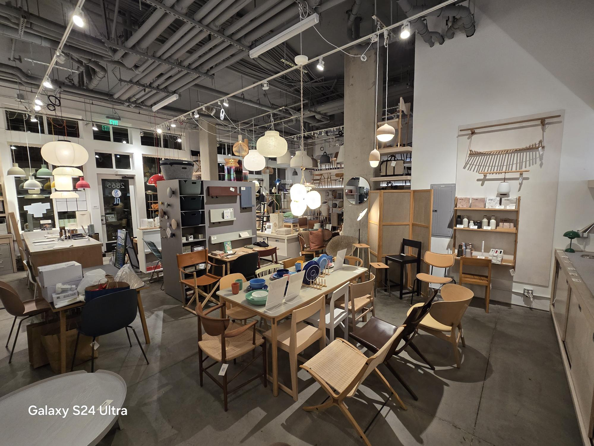 A furniture showroom interior features various wooden chairs, tables, and hanging pendant lights under an industrial ceiling.