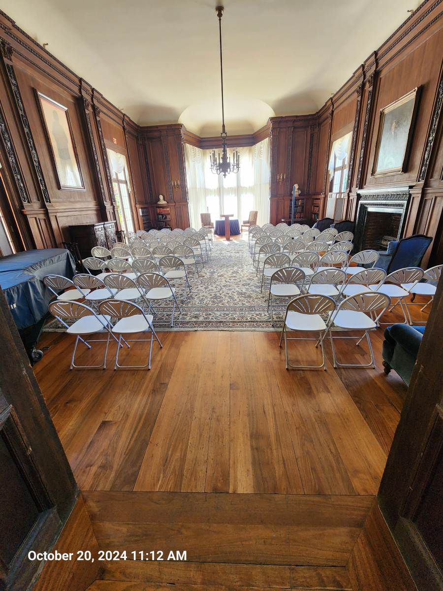 A wooden, historic room set up with rows of folding chairs facing a windowed alcove under a chandelier.