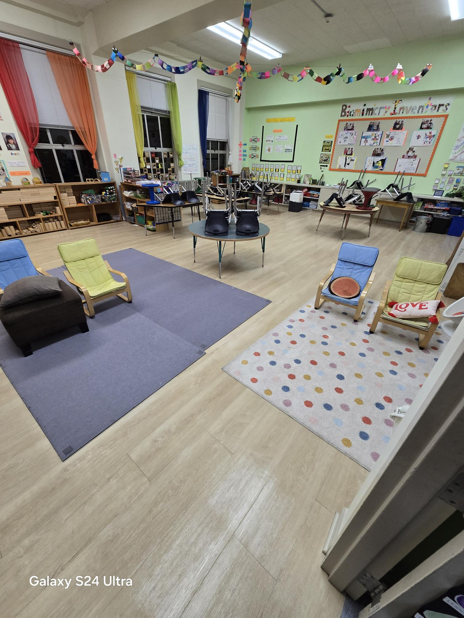 A bright, tidy preschool classroom featuring two floor rugs, small colorful chairs, and children's art on the walls.