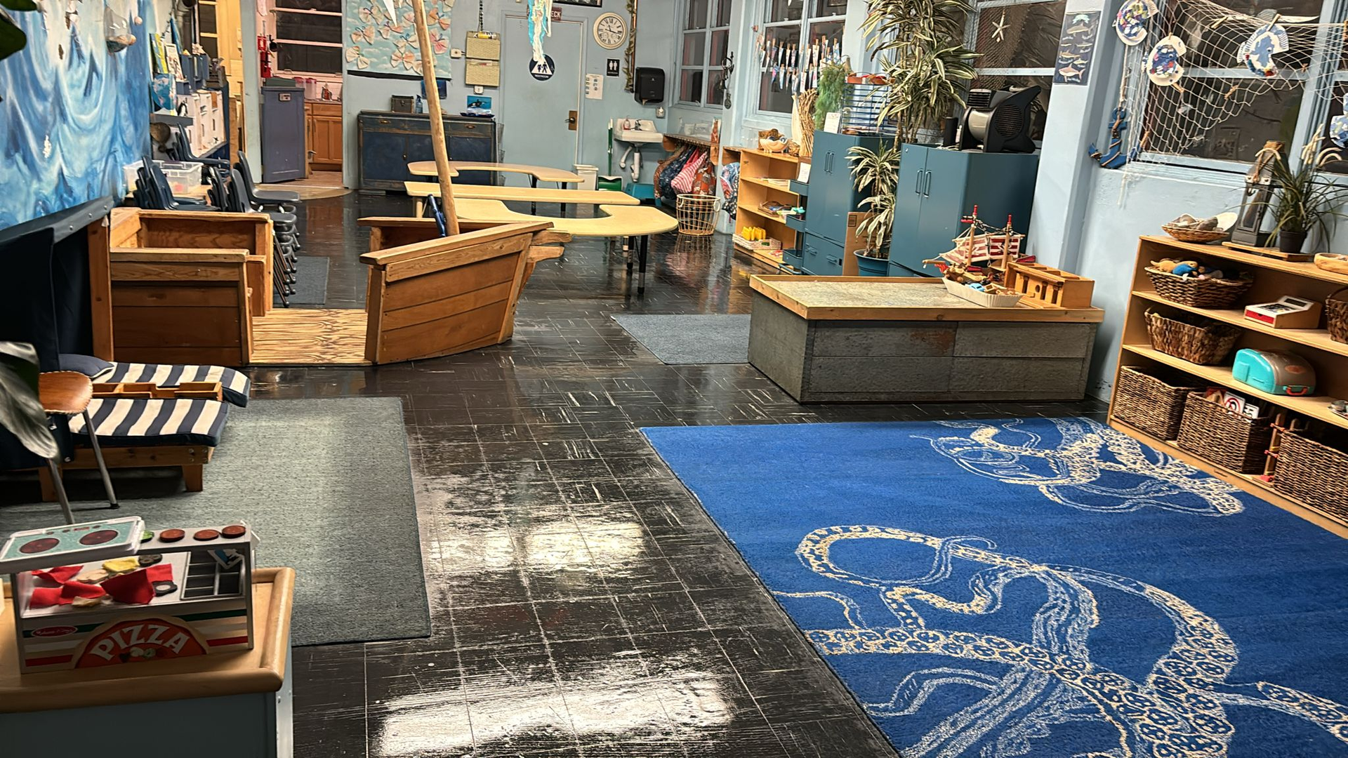 A children's indoor playroom features a blue octopus rug, a wooden play boat, a low table, and shelves with toys.