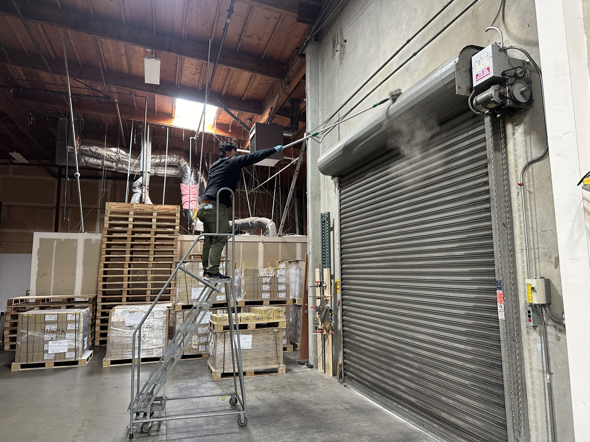 A person stands on a rolling ladder, using a long-handled tool to clean the top of a metal roll-up door in a warehouse.