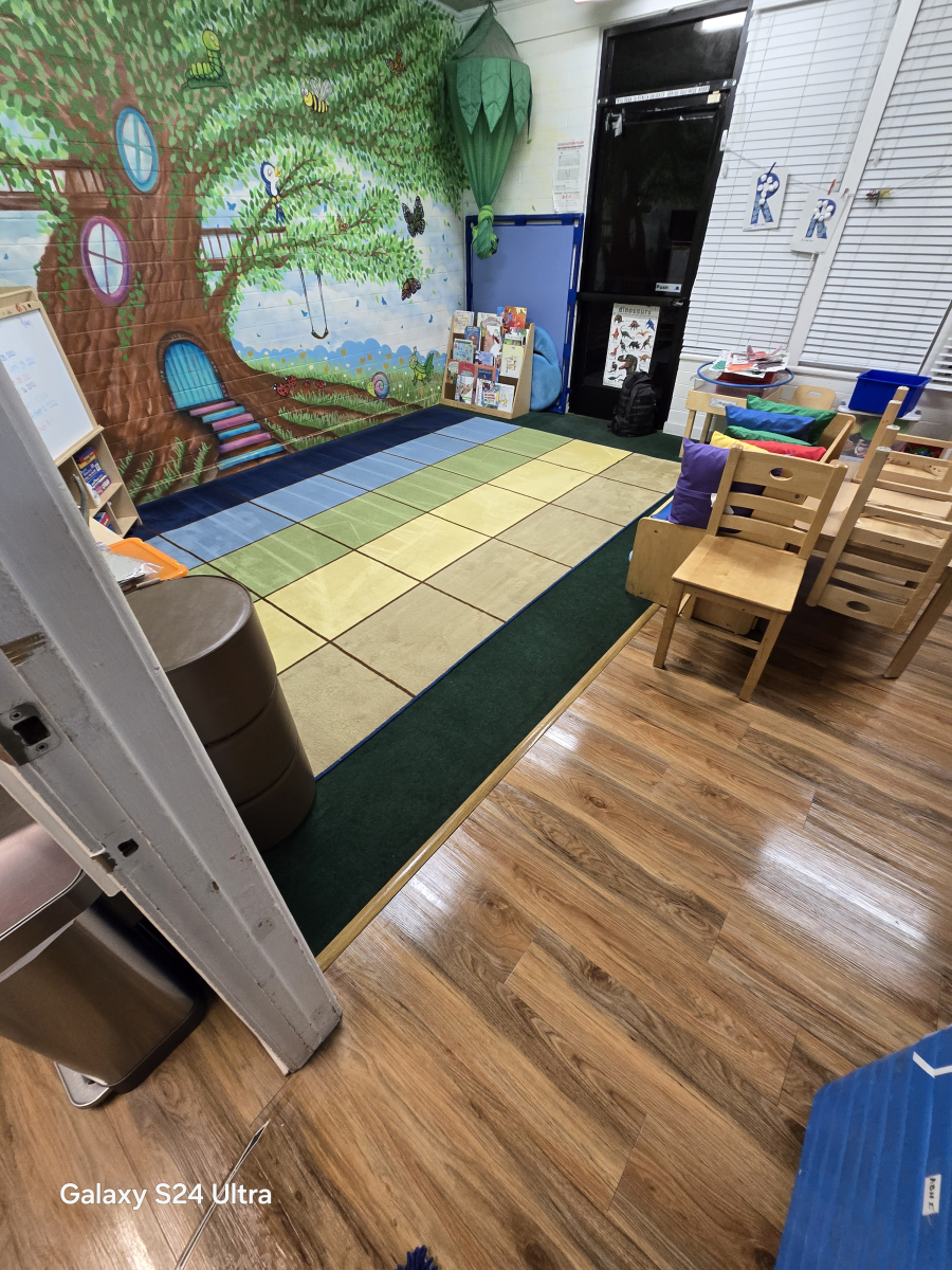 A colorful playroom with a wall-sized tree mural, a square-patterned rug, wooden chairs, and a small bookshelf.