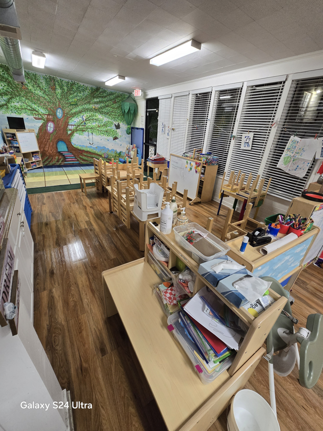 A child’s classroom with a mural of a tree on the wall, wooden floors, organized tables, chairs, and learning supplies.