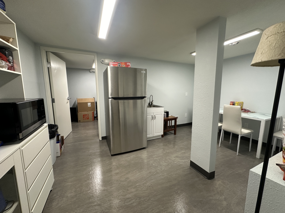 A basement living space with a stainless-steel refrigerator, white cabinets, a table with chairs, and a floor lamp.