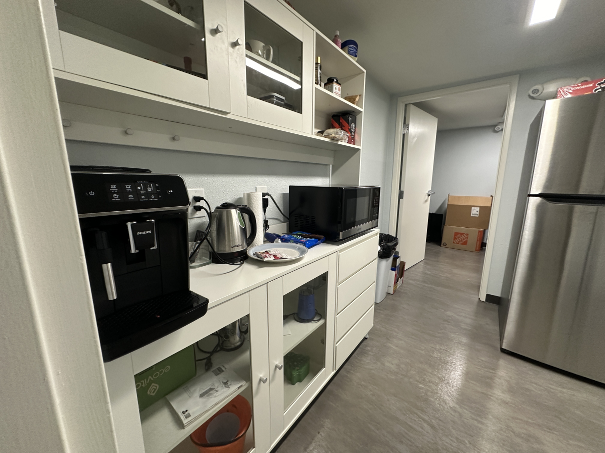 A kitchen area with a white hutch containing a coffee maker, kettle, and microwave, next to a stainless steel refrigerator.